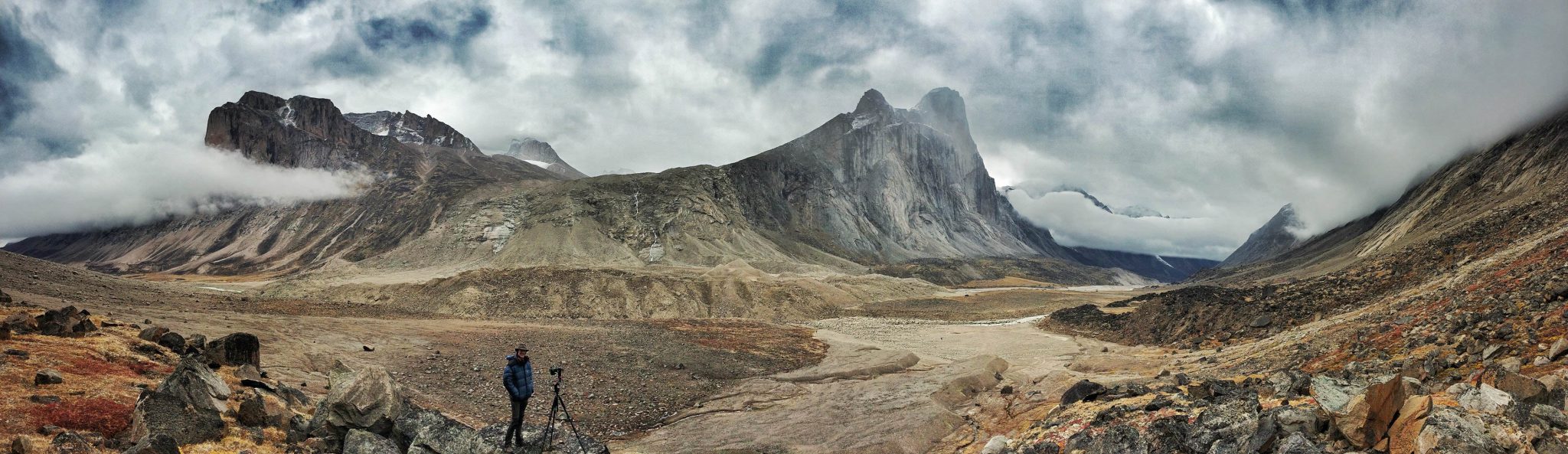 Clear Angle crewmember standing in a valley in front of rocky mountain range and a dramatic, cloudy sky in Baffin Island, Canada. He is standing with a camera and roundshot set up for texture photography.