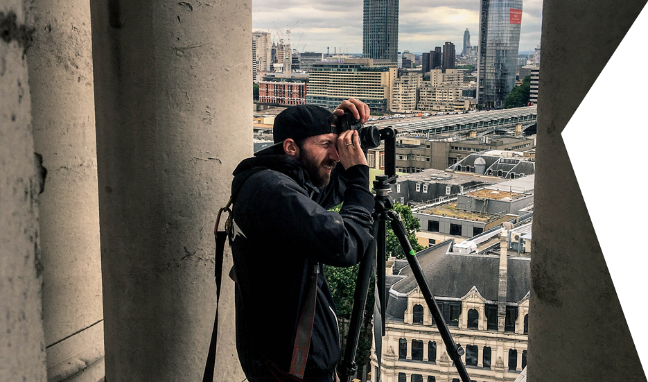 Ross shooting on a rooftop. Clear Angle crewmember looking through the viewfinder of a camera whilst capturing an HDRI from between two stone columns with a city in the background