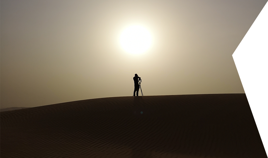 Clear Angle crewmember silhouetted against the sun oon top of a sand dune, setting up a Roundshot to capture an HDRI image.