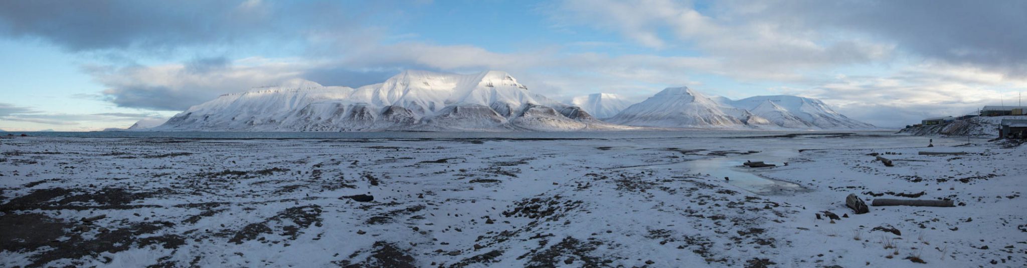 Image from Svalbard in Norway. A snowy plain with black rocks showing through is in the foreground and there's a snowy mountain range in the background.